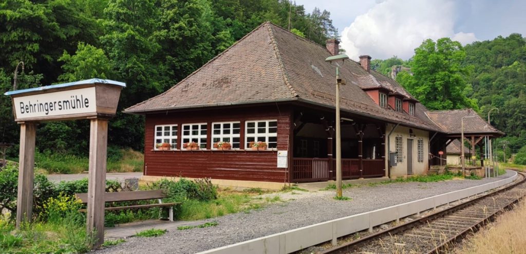 Ansicht der holzgetafelten Wartehalle des Bahnhofes Behringersmühle von außen, mit dem Bahnhofsschild das ein paar Meter davor steht. Bild: W. Bischof